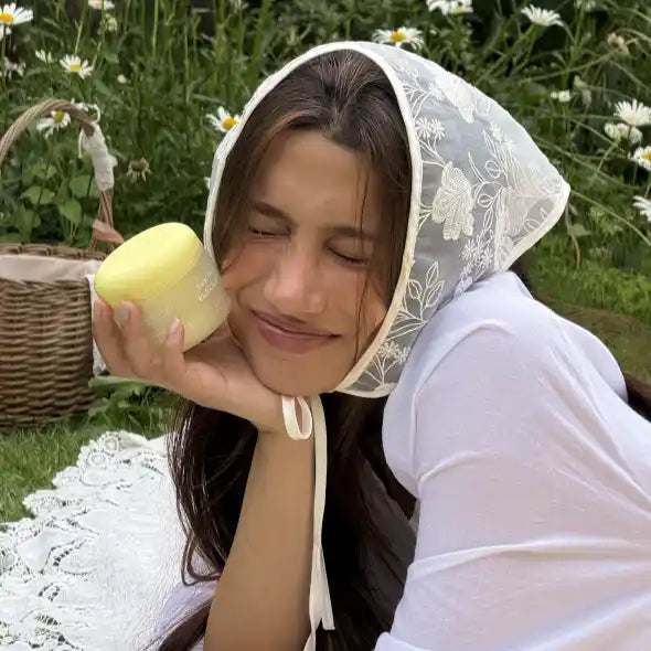 Model in an embroidered headscarf holding the Axis-Y Kojic Yuzu cleanser jar in a sunny daisy field.