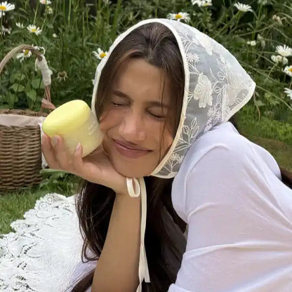Model in an embroidered headscarf holding the Axis-Y Kojic Yuzu cleanser jar in a sunny daisy field.