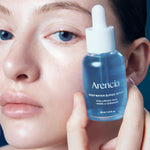 Close-up of a model with glowing, dewy skin holding the Arencia Deep Water Surge Serum bottle near her face.