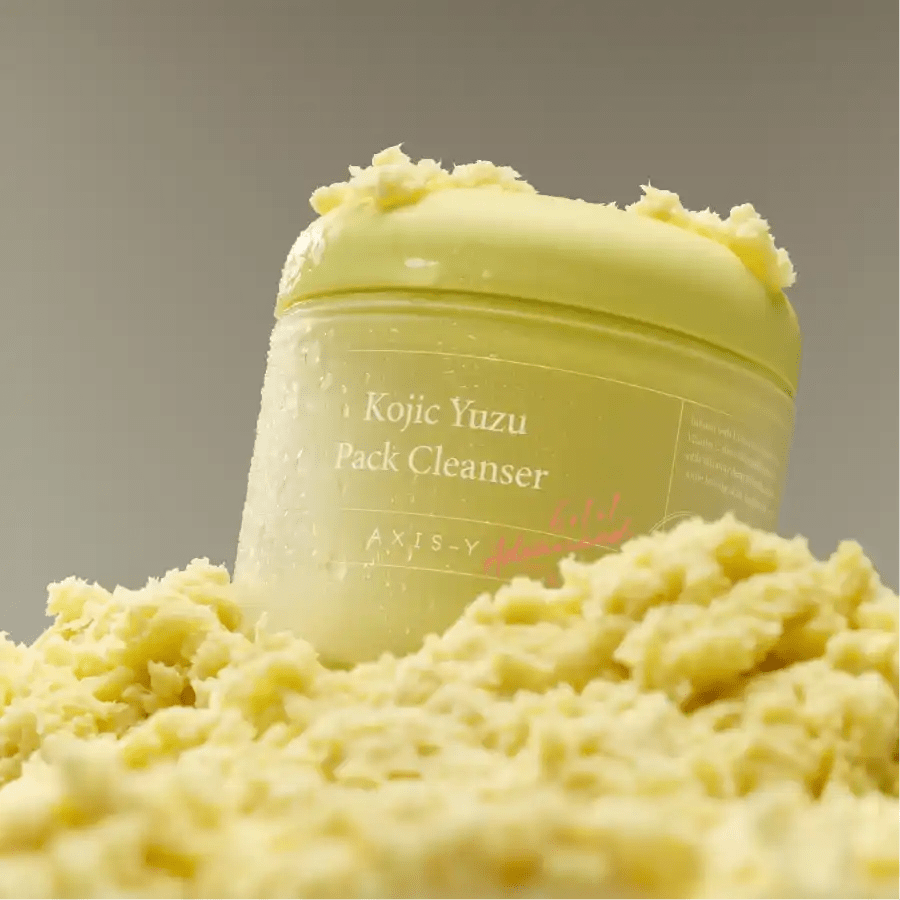 Axis-Y Kojic Yuzu cleanser jar resting on a fluffy, light-yellow soufflé texture in clear lighting.