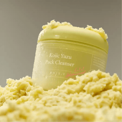 Axis-Y Kojic Yuzu cleanser jar resting on a fluffy, light-yellow soufflé texture in clear lighting.