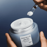 A hand using a clear spatula to scoop Skin1004 Hyalu-Teca Firming Cream from the blue frosted jar.