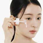 A woman applying the white milky texture of Skin1004 Matrixyl Ampoule to her cheek using a dropper.